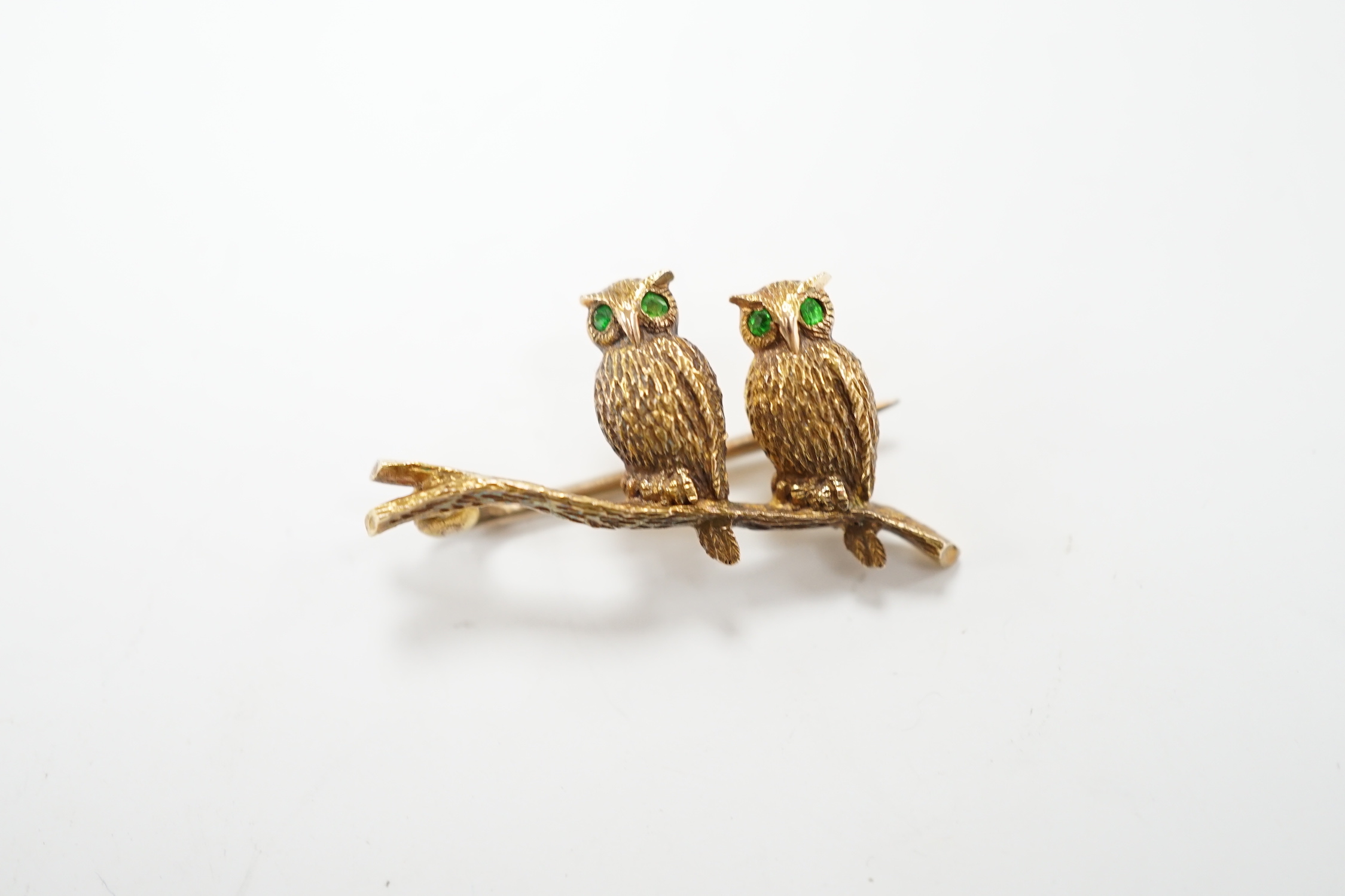 An Edwardian 15ct novelty brooch, modelled as two owls perched on a branch, with four stone green garnet set eyes, 26mm, gross weight 4.1 grams.